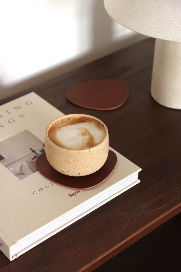 Brauner, asymmetrischer Leder-Untersetzer aus pflanzlich gegerbtem Rindsleder auf dunkler Holzkonsole, mit Cappuccino im Becher