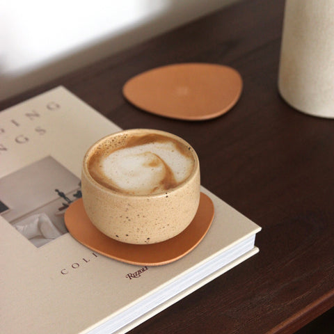 Naturfarbener asymmetrischer Leder-Untersetzer aus pflanzlich gegerbtem Rindsleder auf dunkelbrauner Holzkonsole, mit Cappuccino-Becher