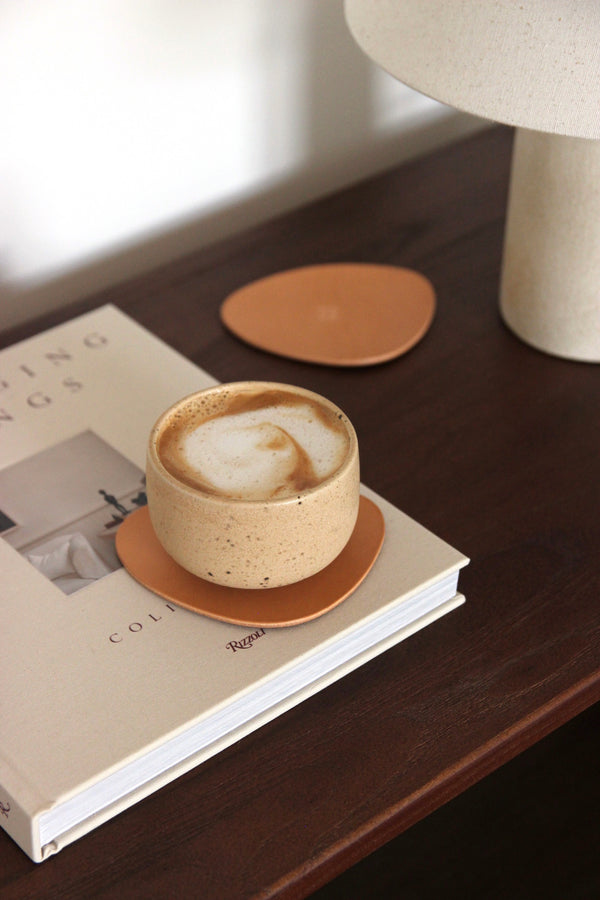 Naturfarbener asymmetrischer Leder-Untersetzer aus pflanzlich gegerbtem Rindsleder auf dunkelbrauner Holzkonsole, mit Cappuccino-Becher