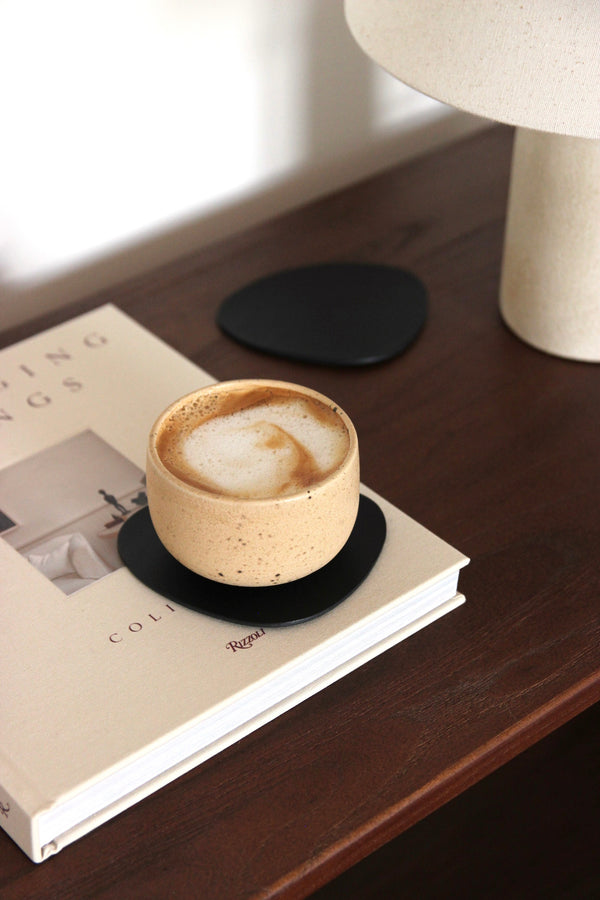 Schwarzer, asymmetrischer Leder-Untersetzer aus pflanzlich gegerbtem Rindsleder auf dunkler Holzkonsole, mit Cappuccino im Keramikbecher